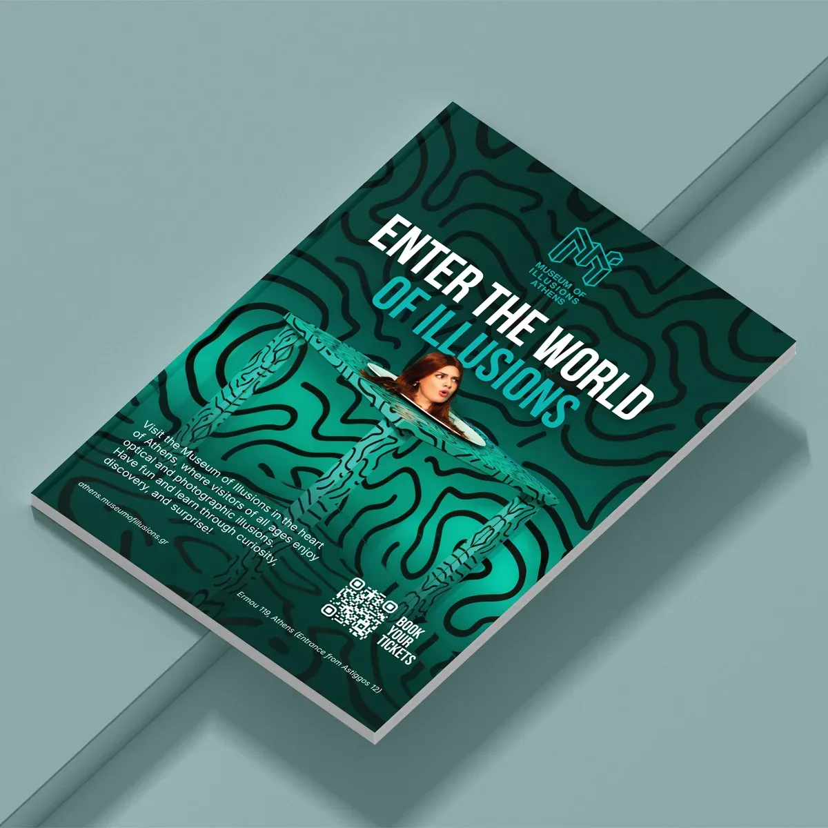 A magazine with the Enter the World of Illusions ad on its back cover: a woman's head on a patterned table, with a Book your tickets QR code